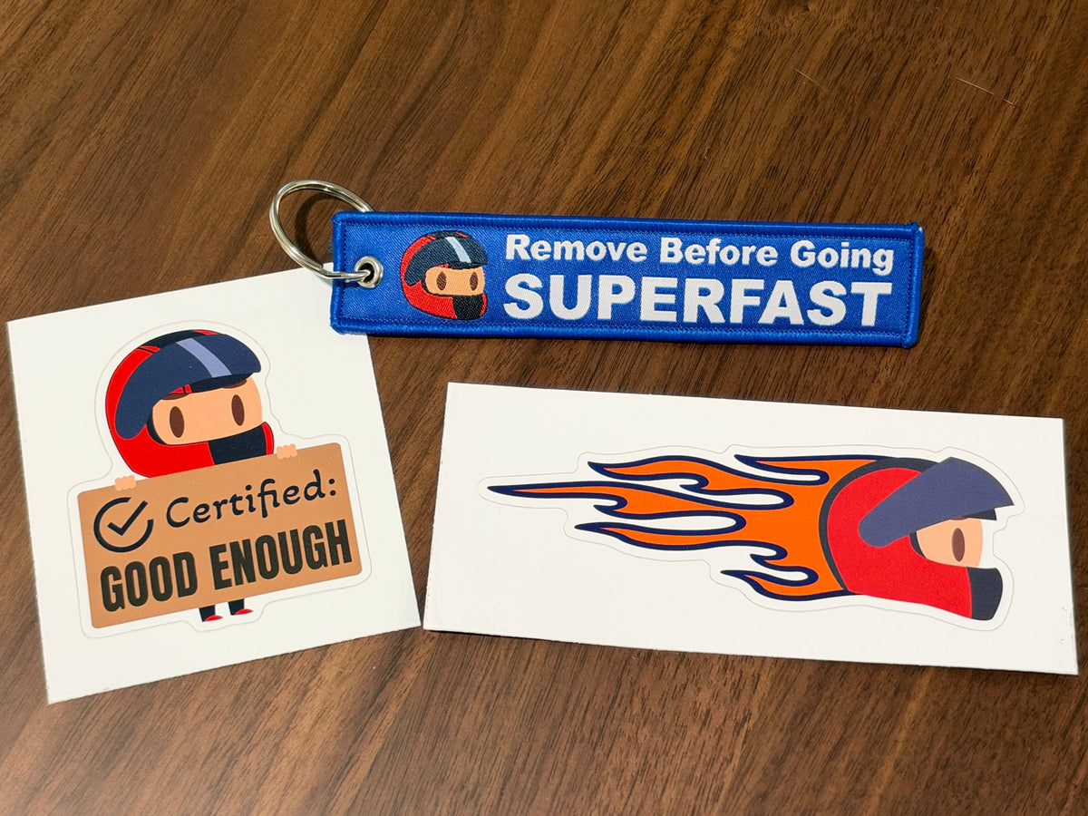 Two racing-themed stickers and a matching keychain lying on a brown wooden table. The keychain is blue with white text reading 'Remove Before Going SUPERFAST' and features a small helmet graphic.
