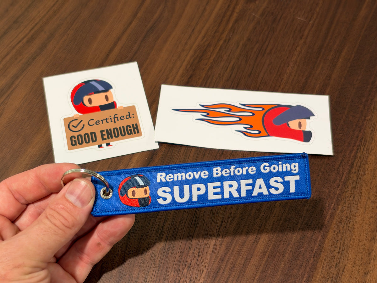 A hand holds a blue embroidered keychain strap with white text reading 'Remove Before Going SUPERFAST' and a small racing helmet graphic. Two small srickers are placed on a wooden table behind it, one reading 'Certified Good Enough'